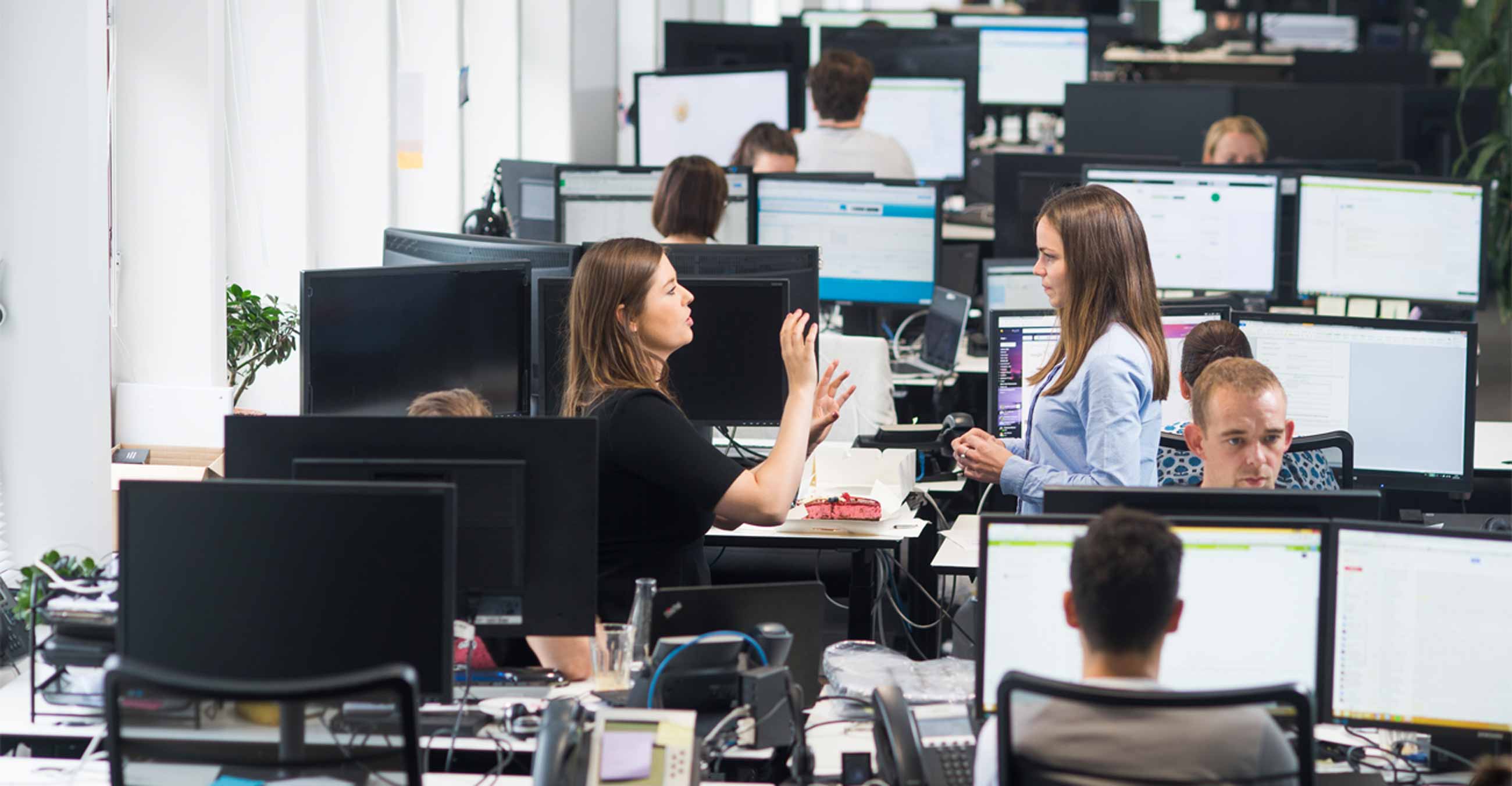 A photograph of two employees engaged in conversation in the midst of desks in Trustpilot's office.